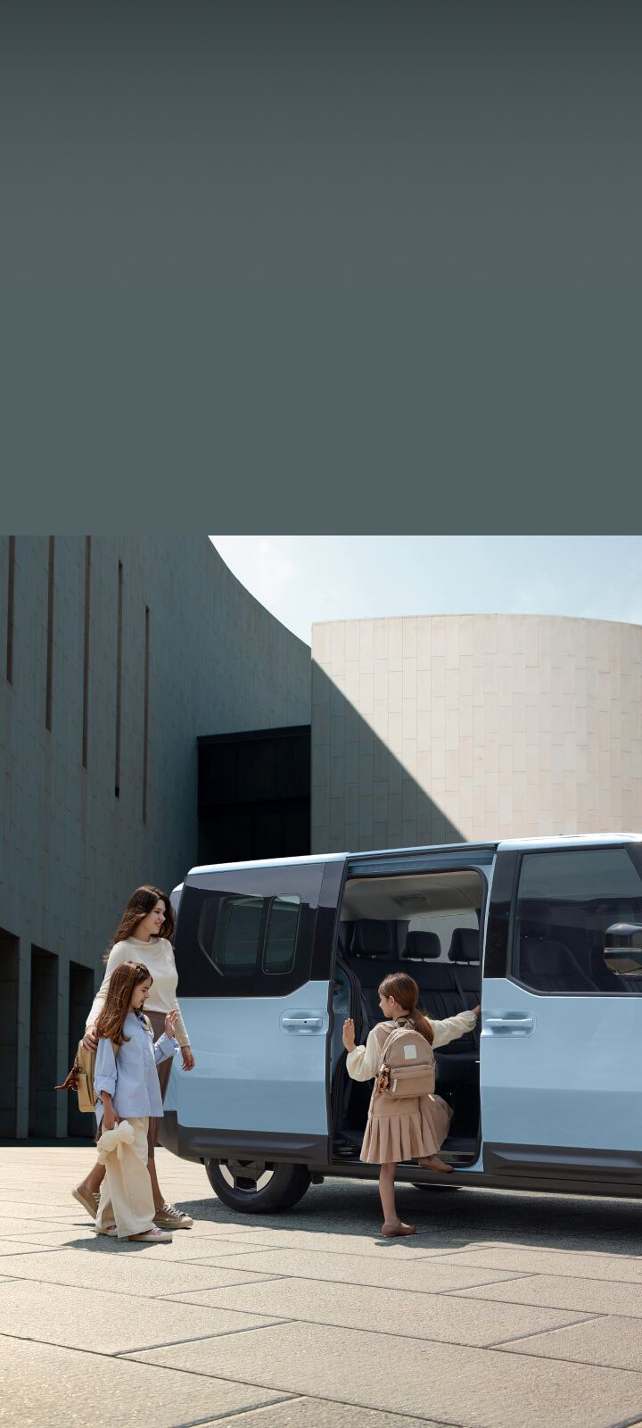 Die seitlichen Schiebetüren des Kia PV5 Passenger öffnen sich, während eine Mutter und ihre Kinder zum Einsteigen bereitstehen.